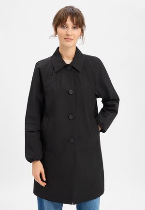 Woman wearing a black buttoned coat with long sleeves and collar, standing against a plain light background.