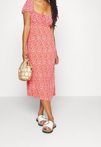 Robe midi rouge à fleurs avec manches courtes bouffantes, encolure carrée et détails élastiques ; accompagnée de sandales blanches et d'un sac tissé.