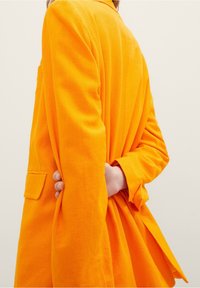 Svetle oranžový blazer s upraveným střihem, obsahující jednu náprsní kapsu a plisovaný zadní design. Vyrobeno z texturované tkaniny.