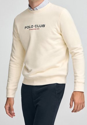 Mężczyzna w kremowej bluzie z napisem „POLO CLUB Established 2012”, w połączeniu z granatowymi spodniami i koszulą w paski z kołnierzykiem.