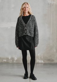 Czarny i szary melanżowy kardigan z dzianiny o luźnym kroju i zaokrąglonym dole, zapinany na guziki, noszony na czarnej sukience i rajstopach. Do kompletu czarne buty.