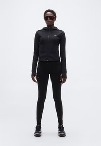 Αθλητικό άτομο που φοράει μαύρο ζιπ-άπ μπουφάν, μαύρο κολάν, αθλητικά παπούτσια και μεγάλα κόκκινα αθλητικά γυαλιά ηλίου, στέκεται μπροστά σε απλό φόντο.