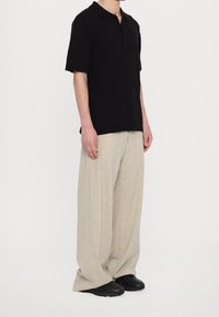 Henkilö, joka pukeutuu mustaan lyhythihaiseen kauluspaidastaan, vaaleisiin beigeihin housuihin, jotka peittävät mustat kengät, seisoo tasaisella taustalla.