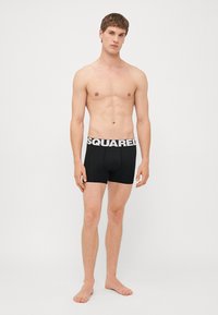 Černé trenýrky s bílým logem "SQUARED" na pásku. Tkanina vypadá hladce a elasticky, navržena pro pohodlí.