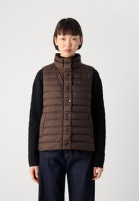 Kvinne som har på seg en brun quiltet puffer vest med trykknapper over en svart kabelstrikket genser og mørke jeans, står mot en hvit bakgrunn.