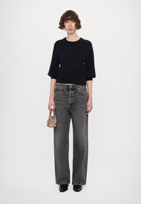 Schwarzer Strick-Crop-Pullover, lockere graue weit geschnittene Jeans, kleine gemusterte Tasche und schwarze Schuhe. Einfache Silhouette mit Texturkontrast.