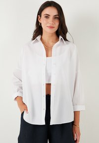 Camicia bianca con bottone, dal fit rilassato, maniche lunghe arrotolate ai polsini, tessuto traslucido e orlo dritto. Indossata sopra un top corto.