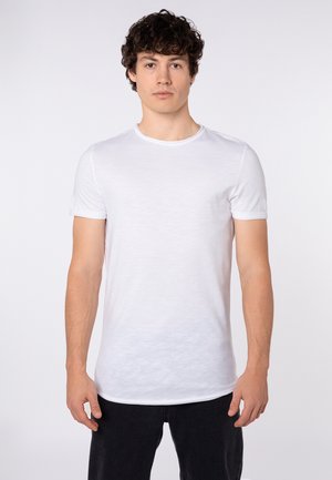 Junger Mann mit lockigem dunklem Haar, der ein schlichtes weißes kurzärmeliges T-Shirt und schwarze Hosen trägt, steht vor einem schlichten hellen Hintergrund.