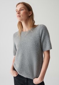 Lichtgrijze, korte mouwen woltrui met een zachte, pluizige textuur en een ronde halslijn, gecombineerd met donkere broek, handen in de zakken.