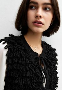 Zwarte gebreide vest met een textuur van lusfranje. Vastgemaakt met een strikje bij de hals, gelaagd over een witte blouse, met subtiele sieradenaccenten.