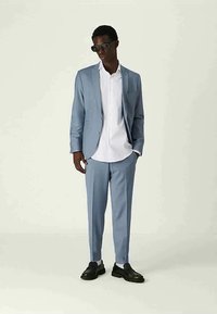 Mies, joka on pukeutunut vaaleansiniseen pukuun valkoisen paidan, mustien loafereiden ja tummien aurinkolasien kanssa, seisoo tasaisella vaalealla taustalla.