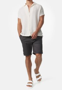 Chemise blanche à manches courtes avec boutons, tissu texturé ; short roulé gris foncé ; sandales blanches avec bride à boucle ; tenue décontractée.