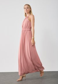 Maxiklänning i mjuk rosa tyg med ett samlat elastiskt midjeband och halterneck. Har en svängd kjol och strappy klackar.