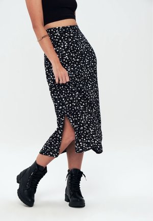 Femme portant une jupe midi noire à motif floral avec une fente latérale, un crop top noir, des bottines noires à lacets et un bracelet jonc doré.
