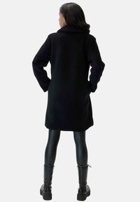 Black fleece coat with a collar, mid-length design, dropped shoulders, side pockets, worn over shiny black leggings and chunky black boots.