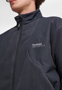 Mörkgrå jacka med en mjuk fleeceyta, släta tygsektioner, hög krage och framzip. Logotypen tryckt på bröstet.