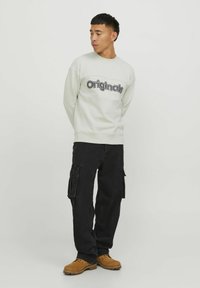 Sudadera de cuello redondo gris claro con "originales" impreso en texto negro en negrita, combinada con pantalones cargo negros y zapatos de trabajo color marrón.