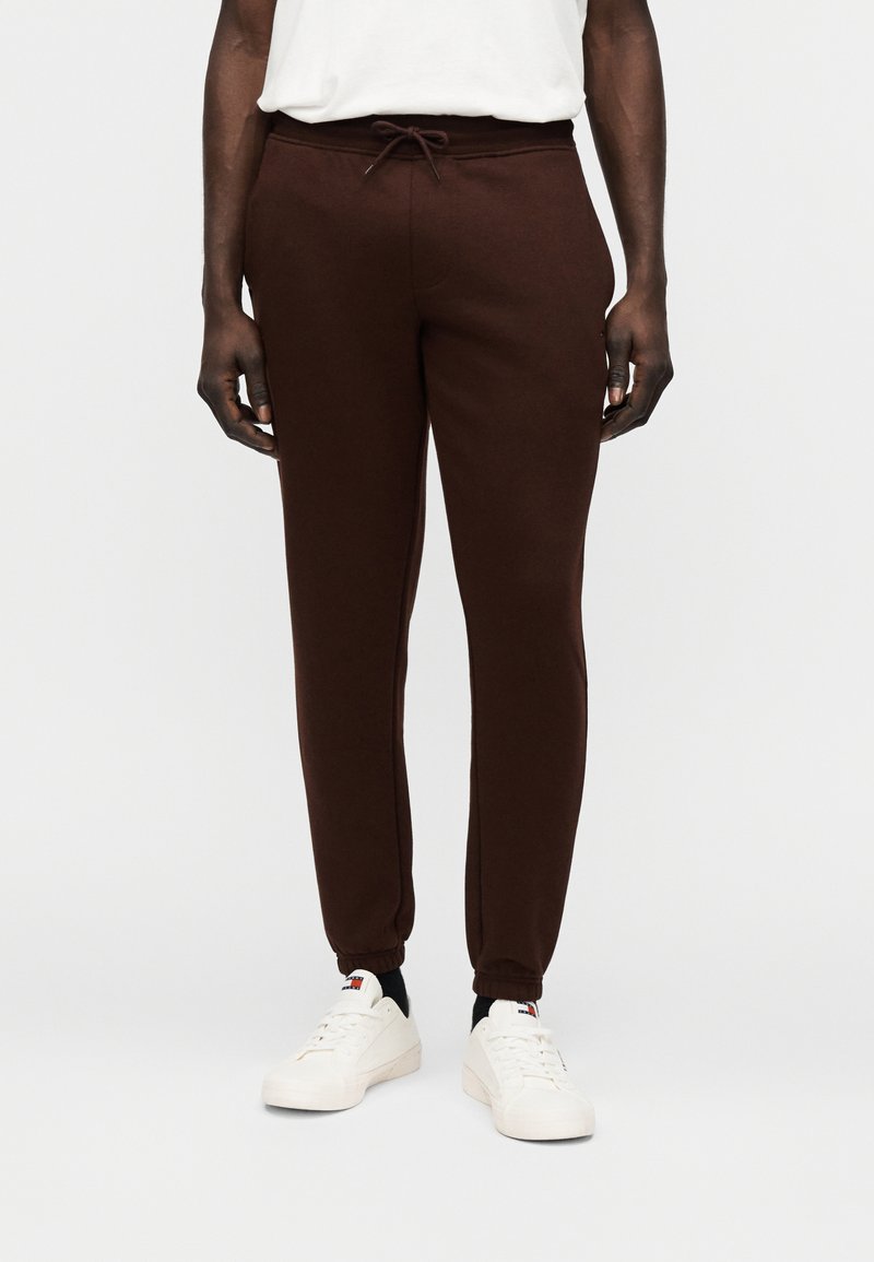 Καφέ φόρμα από απαλό ύφασμα, με ελαστική μέση και κορδόνι, εφαρμοστές μανσέτες και πλαϊνές τσέπες. Συνδυασμένο με λευκά αθλητικά παπούτσια.