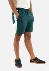 Calções verdes e brancos com cintura elástica e cordão; design de frente plana; combinados com sapatos pretos com detalhes em laranja.