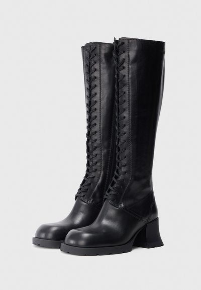 Botas de cuero negras hasta la rodilla con una textura suave, cierre frontal con cordones y tacón grueso. Cuentan con una cremallera lateral para facilitar su uso.