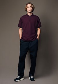 Polo en bordeaux avec un col zippé, associé à un pantalon noir marine et des baskets noires avec des lacets blancs, se tenant devant un fond neutre.