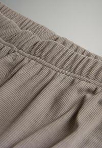 Ljust brunt texturerat tyg med ribbat mönster, elastisk midja och mjuka draperingar. Närbild visar materialdetaljer och design.