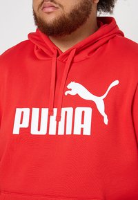 Sudadera con capucha roja y bolsillo frontal, que presenta un gran logo blanco de "PUMA" y la silueta de un gato saltando. Tejido suave y texturizado.