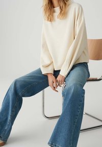 Personne aux cheveux longs assise sur une chaise en bois, portant un pull crème oversize et un jean bleu à jambes larges sur fond uni.