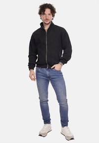 Giacca nera con zip e finitura testurizzata, abbinata a jeans in denim blu e sneakers bianche, indossata da una persona in piedi.