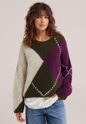 Vrouw met golvend haar draagt een meerkleurige argyle trui in crème, donkergroen en paars over een wit topje en blauwe spijkerbroek.