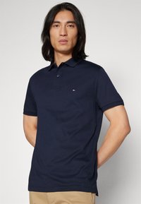 Navy blauwe polo met korte mouwen, een kraag, een knoopsluiting met twee knopen en een klein logo op de borst. Katoenen stof met een gladde textuur.
