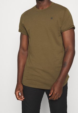 Homme portant un t-shirt vert olive à manches courtes avec un petit logo noir sur la poitrine, associé à un pantalon noir, debout devant un fond uni.