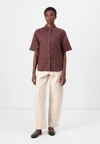 Chemise à manches courtes en lin brun, associée à un pantalon large crème, portée avec des ballerines noires. Design simple avec un col classique.