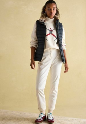 Schwarze Steppweste mit Pelzkragen über einem cremefarbenen Pullover mit rot-schwarzem Muster, kombiniert mit cremefarbenen Hosen und burgunderroten Turnschuhen mit blauen Akzenten.