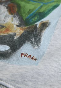 Koszulka z nadrukiem przedstawiającym kolorową ilustrację w odcieniach zieleni, pomarańczu i czerni, nadrukowaną na jasnoszarym materiale, podpisaną "FRANK".