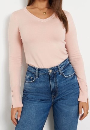 Frau trägt einen hellrosa langärmligen V-Ausschnitt-Pullover mit Knopfdetails an den Manschetten und hochtaillierten blauen Jeans.