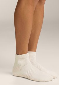 Pár világos bézs színű bordázott bokazokni, amelyet lábakon viselnek egy semleges, egyszínű háttér előtt, bemutatva a zokni textúráját és illeszkedését.