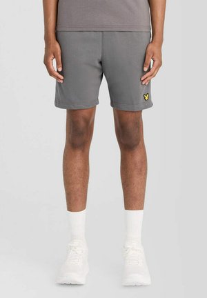 Mann trägt graue Shorts mit gelbem Logo am linken Bein, weiße Sportsocken und weiße Sneaker, steht vor einem schlichten hellen Hintergrund.