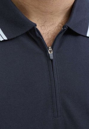 Gros plan d'un polo bleu marine partiellement zippé avec un col laissant entrevoir des poils de poitrine en dessous.