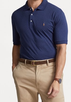 Polo shirt blu navy con colletto a tre bottoni e maniche corte; presenta un piccolo logo ricamato arancione. Abbinato a chino color khaki e una cintura marrone.