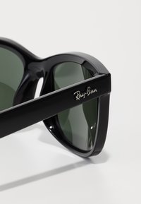 Ochelari de soare din plastic negru cu lentile verzi, având un logo Ray-Ban pe templu și un design elegant, unghiular.