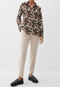 Camicia a maniche lunghe con bottini, caratterizzata da un motivo astratto marrone, crema e nero, abbinata a pantaloni beige a gamba affusolata e mocassini neri.