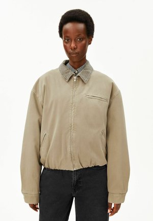 Personne portant une veste beige zippée avec un col en velours côtelé et un pantalon foncé, debout devant un fond blanc uni.