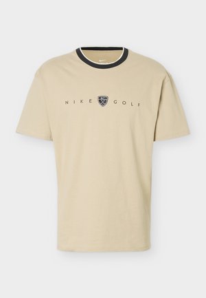 T-shirt de golf Nike beige avec bordure de col noire et logo présentant des clubs de golf croisés à l'intérieur d'un écusson sur la poitrine.