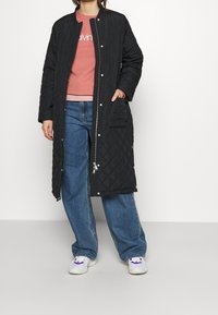 Czarny pikowany długi płaszcz z okrągłym dekoltem, zatrzaskami i kieszenią boczną, noszony na jasnoróżowym swetrze oraz niebieskich spodniach dzwonach.