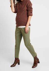 Těhotná osoba oblečená v hnědém pleteném svetru, olivově zelených kalhotách a hnědých kotníkových botách na podpatku, stojící před jednobarevným pozadím.