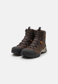 Brązowe buty trekkingowe o solidnym designie, teksturowana gumowa podeszwa, wzmocniony nosek, pomarańczowe akcenty oraz regulowane sznurówki.