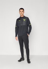 Svart athletic pullover med glidelås i halsen, polstret brystparti og neongule detaljer. Slim-fit svarte bukser og svarte fotballsko fullfører antrekket.