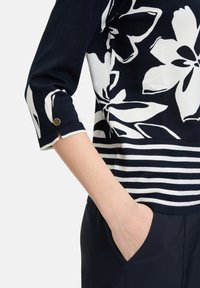 Marineblauwe en witte pullover met bloemenpatroon en gestreepte ondersectie. Driekwartmouwen met knoopdetails. Zachte gebreide stof.
