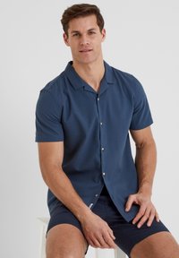 Short-sleeve button-up shirt in textured navy fabric. Features a classic collar and subtle button accents. Worn with navy shorts.